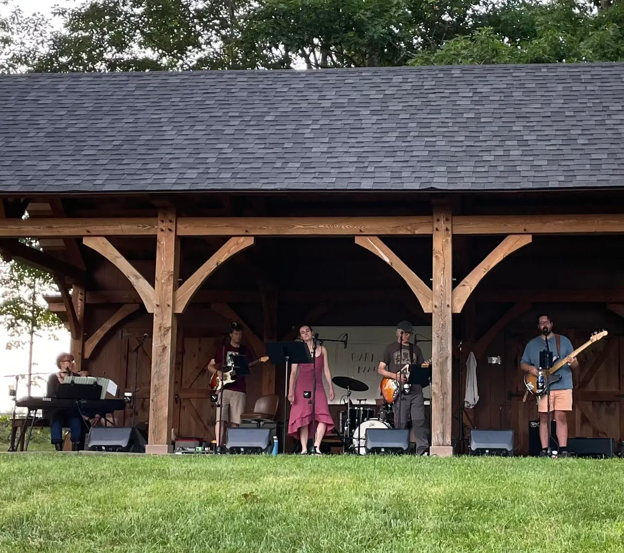The Barn Band playing an open-sided timber pavilion on a summer evening, with a singer in a red dress at the front